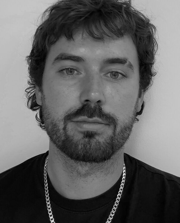A black and white headshot of a man weith an eyebrow slit and silver chain on over a black t shirt