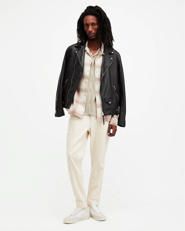 Portrait of a male model wearing a black leather jacket layered over a cuban collar shirt and a checked overshirt, wearing cream trousers and leather trainers.
