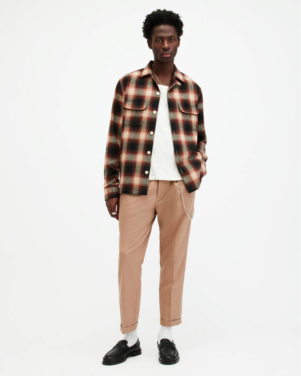 Portrait of a male model wearing a black checked overshirt, a white t-shirt, light brown trousers and black leather loafers.