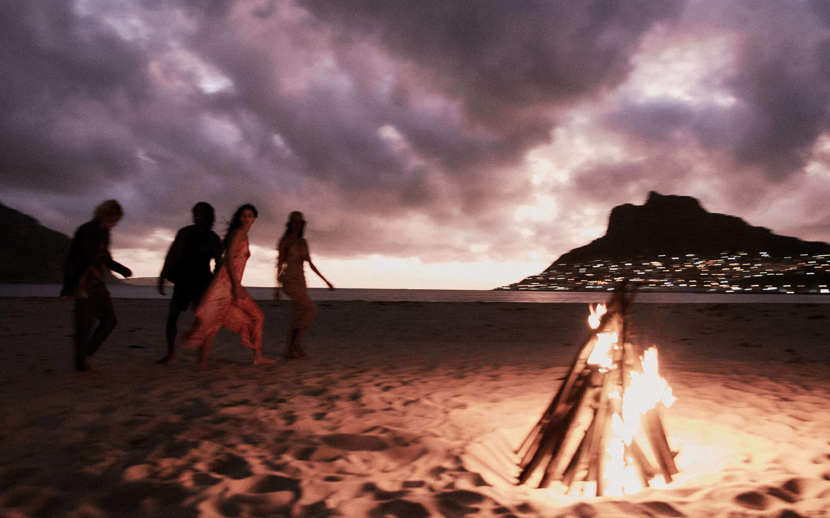 Men and women walking past a bonfire on a beach at night