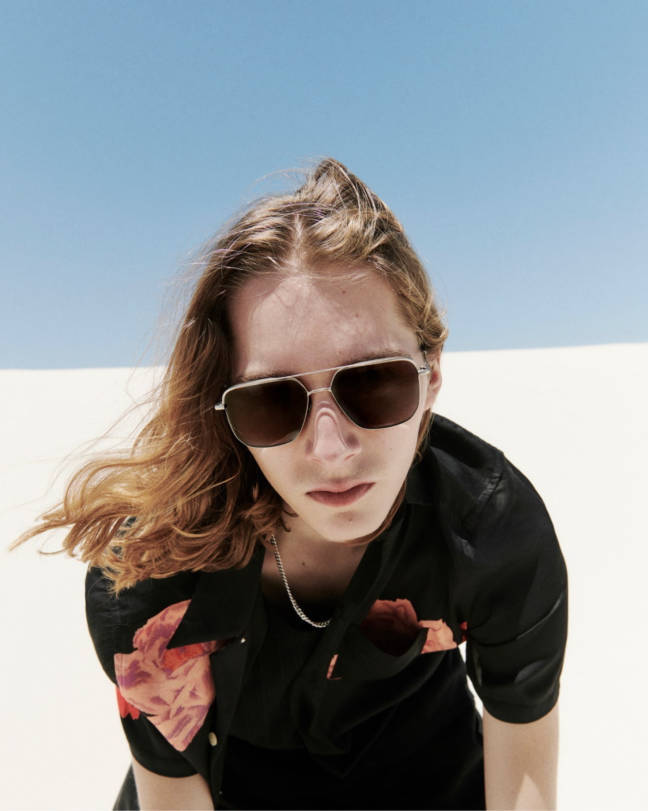 A man wearing aviator sunglasses, a chain necklace and a printed black shirt standing in front of a white dune.
