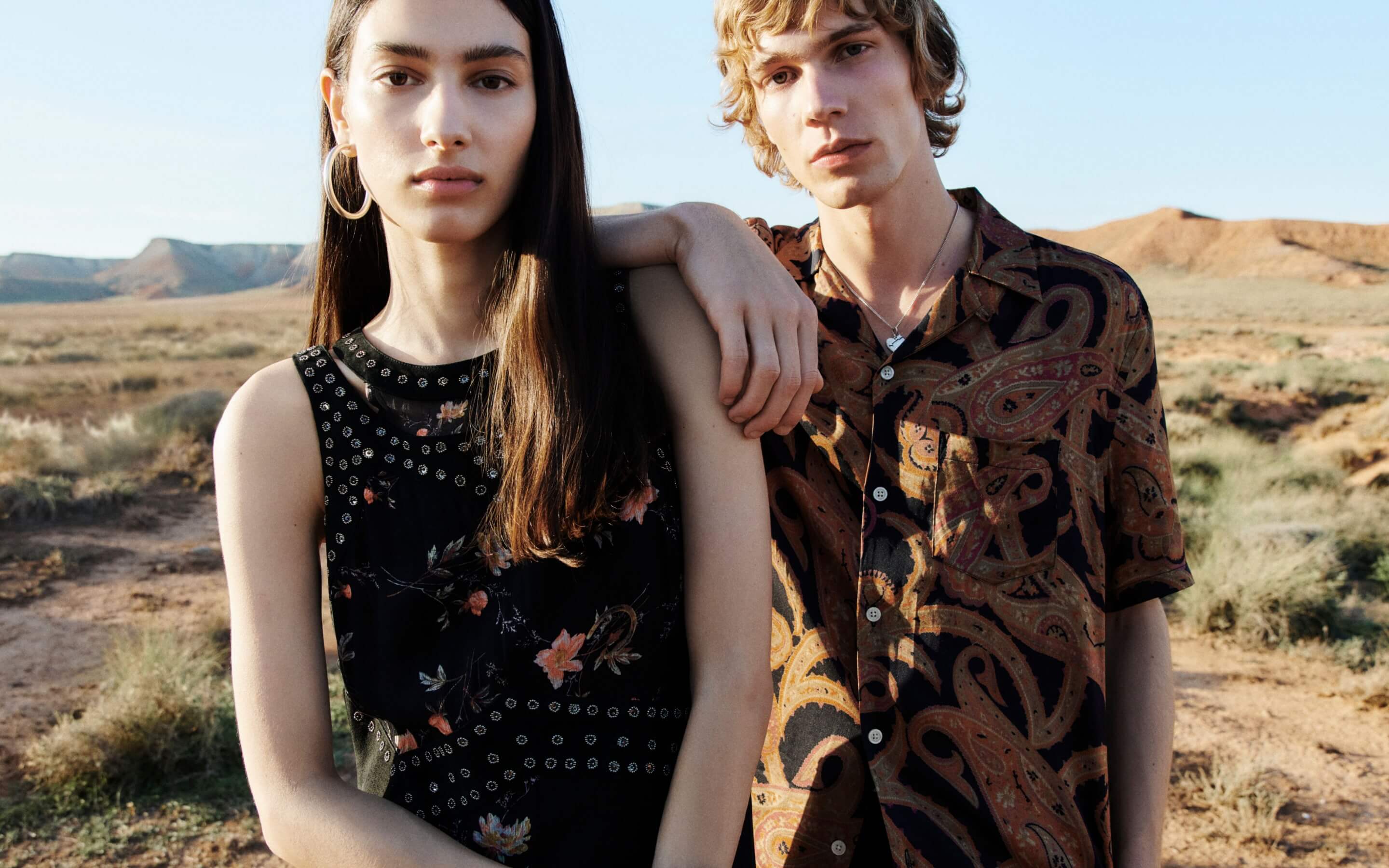 A woman wearing a black embellished floral dress and hoop earrings and a man wearing a paisley print shirt and a heart pendant silver necklace standing in front of a desert landscape.