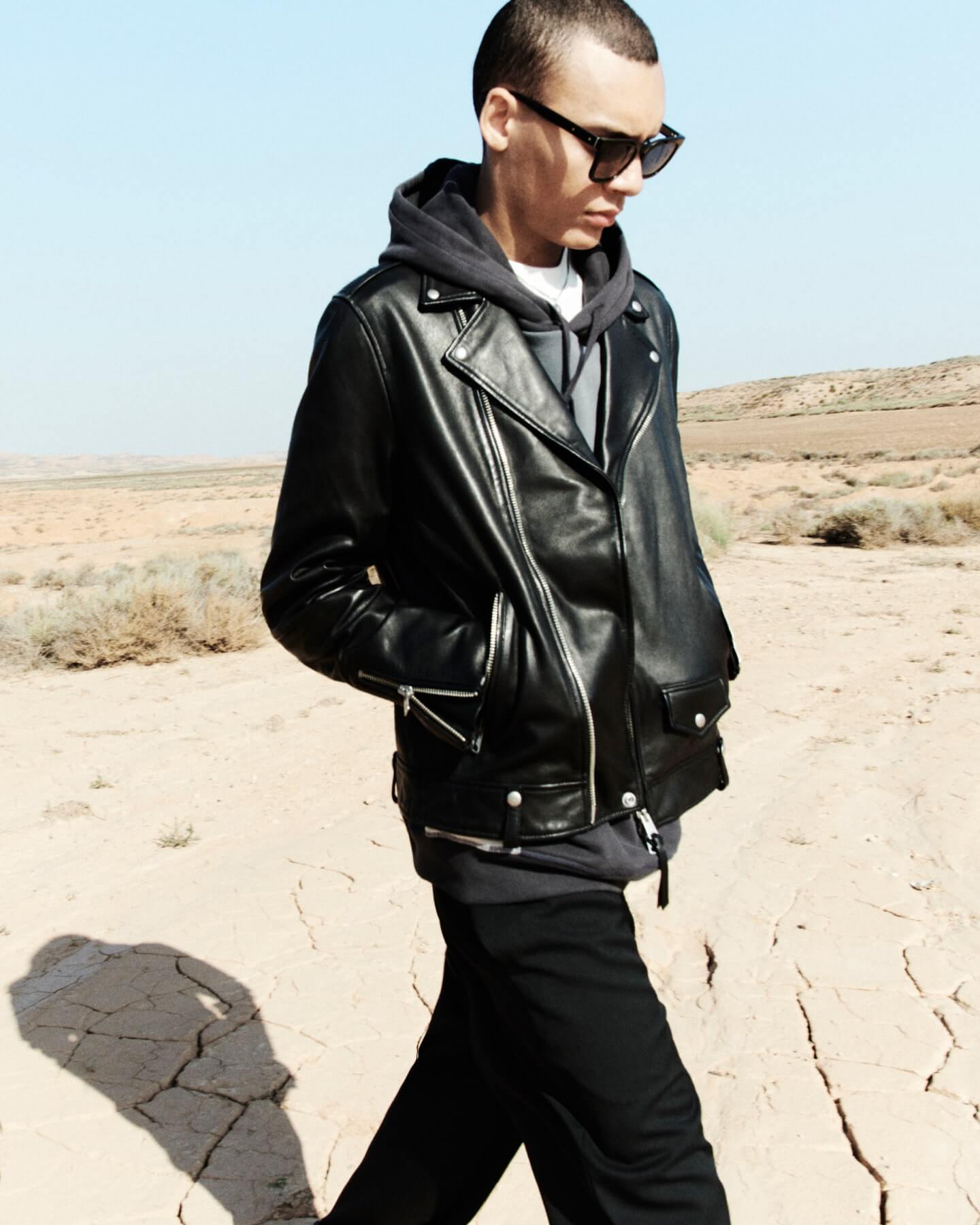 Man wearing a black leather jacket layered over a hoodie and black sunglasses walking in the desert.