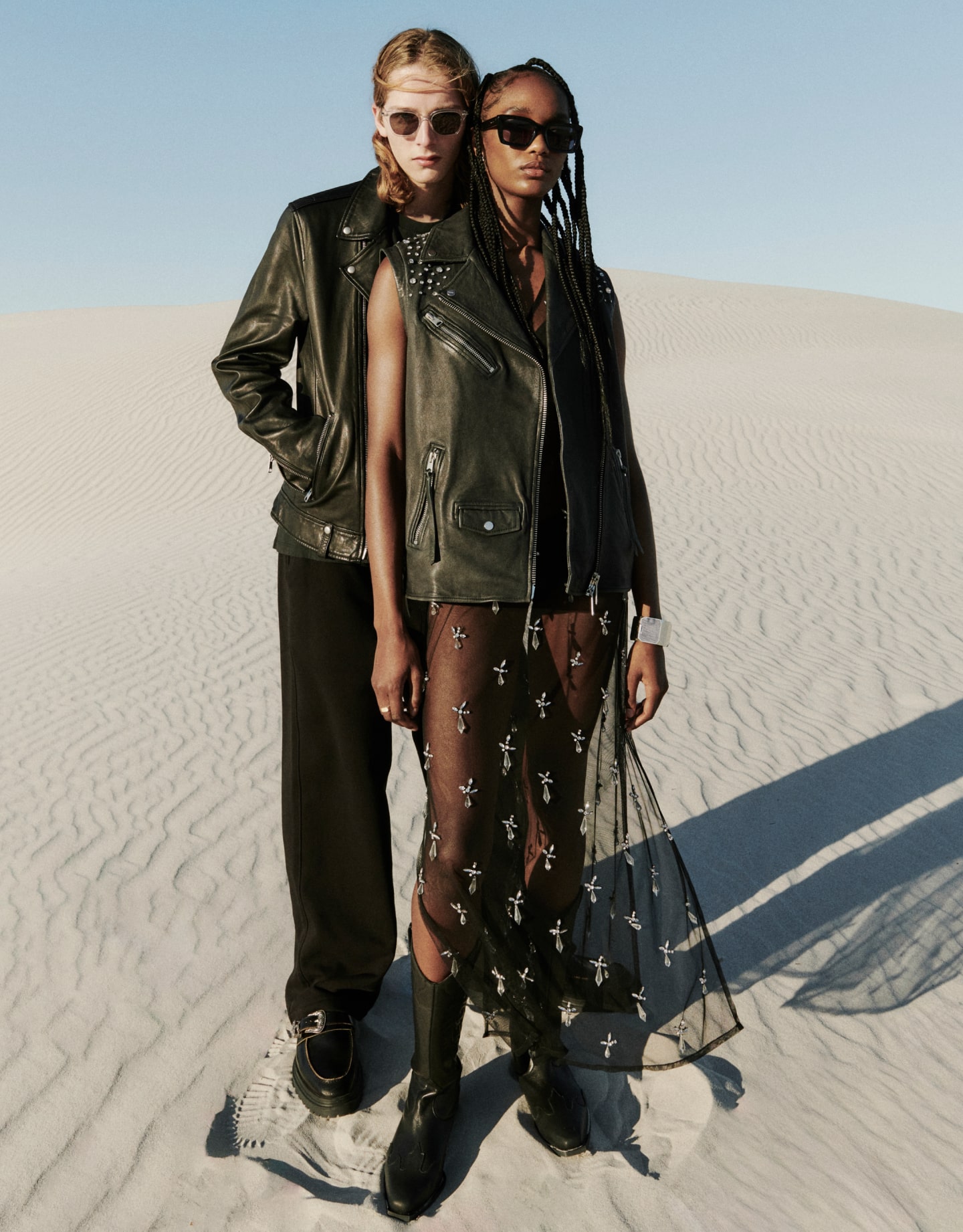A woman wearing a sleeveless black leather jacket and black sunglasses standing in front of a man wearing transparent sunglasses and a black leather jacket standing in the desert.
