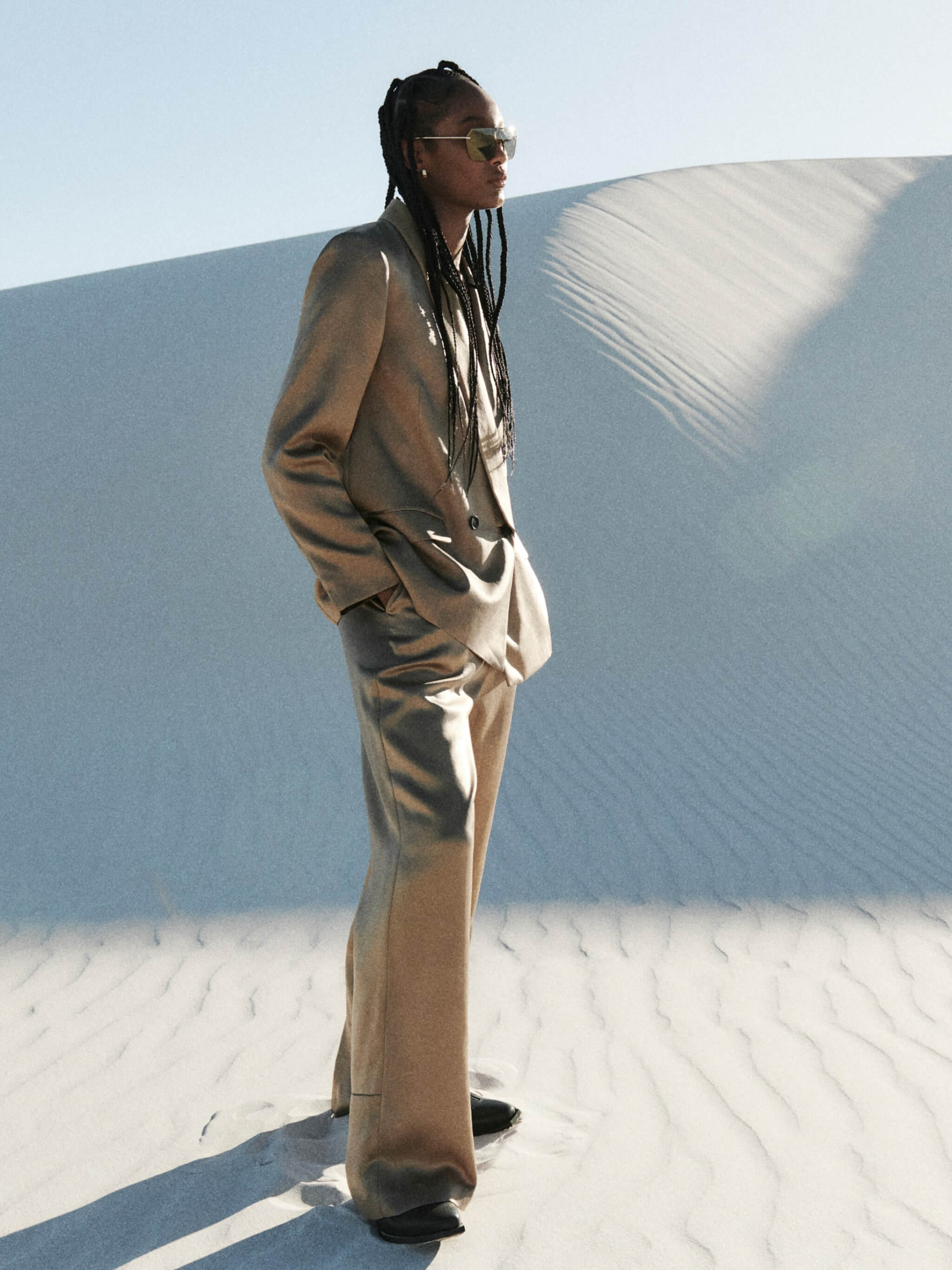 A woman standing in the desert wearing a light brown tailored suit and sunglasses.