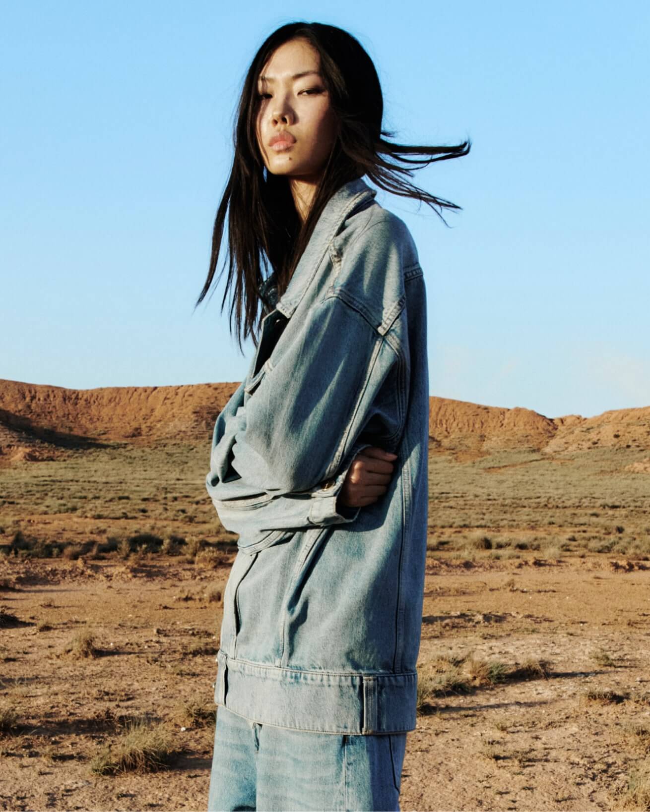 A woman wearing a denim jacket and denim trousers with her hair floating in the wind.