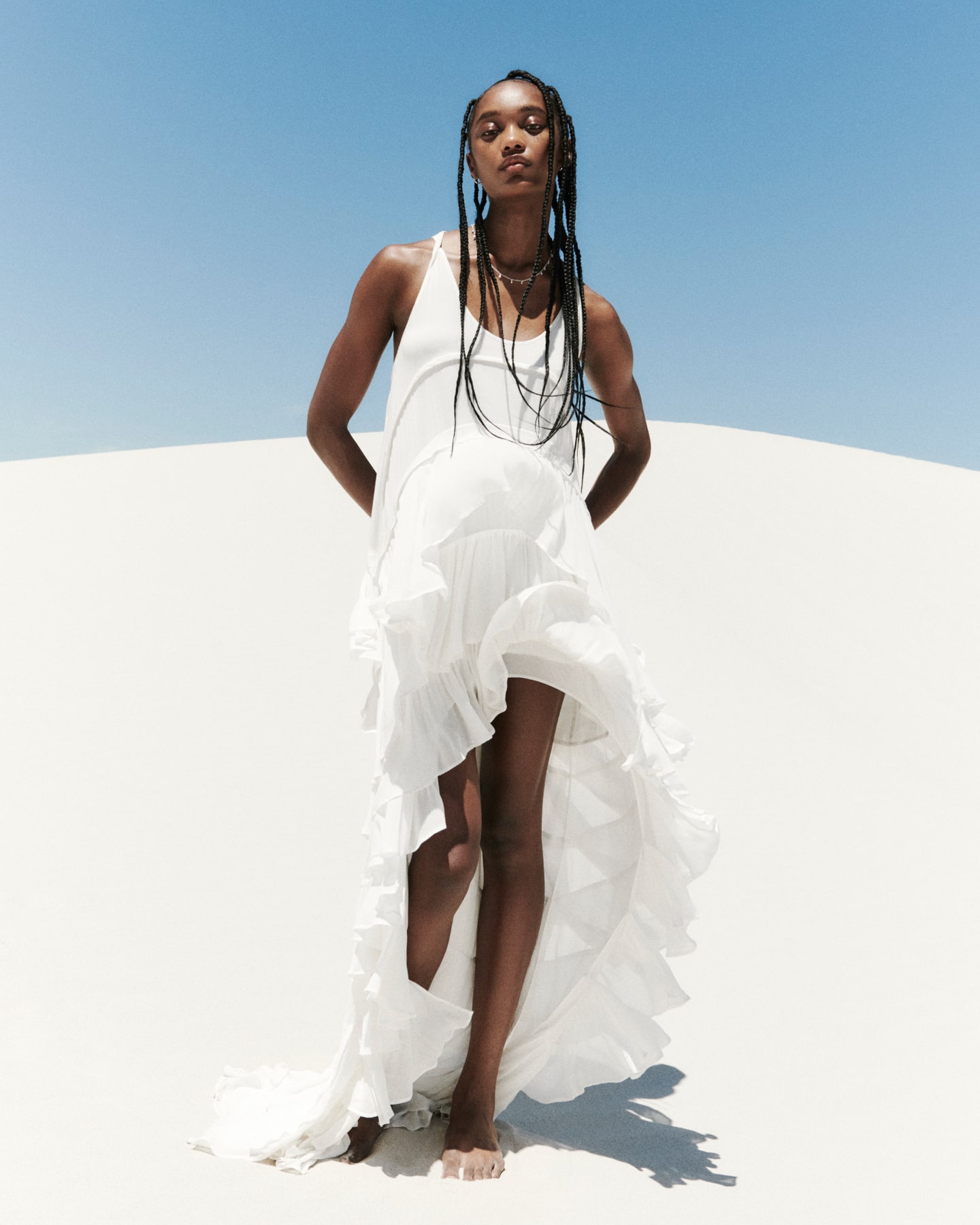 Eine Frau in einem asymmetrischen weißen Kleid steht vor einer weißen Düne.