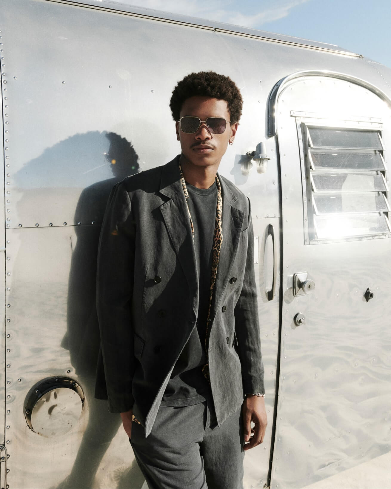 A man wearing a grey tailored suit and sunglasses standing against an old fashioned aeroplane.