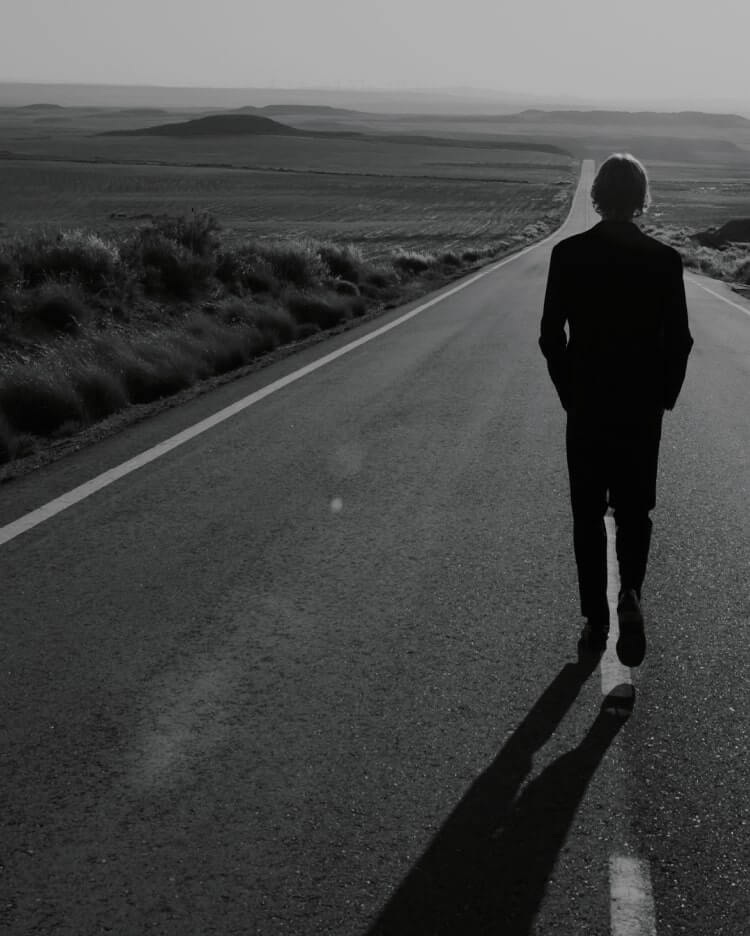Black and white photograph of a man walking along an empty road in the desert.